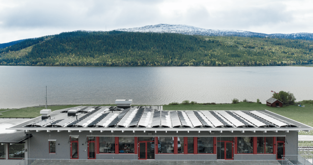 Aurinkopaneeleilla varustettu rakennus veden äärellä, taustalla metsäinen vuoristo.