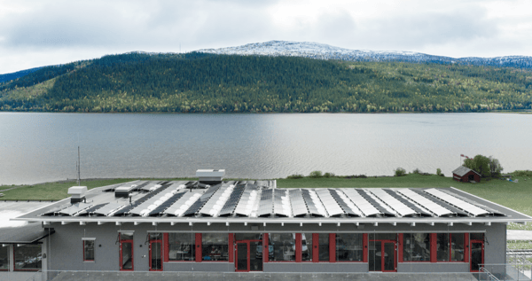 Aurinkopaneeleilla varustettu rakennus veden äärellä, taustalla metsäinen vuoristo.
