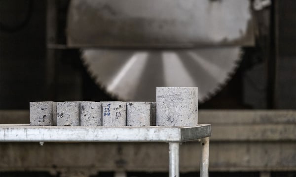Lieriömäisiä betoninäytteitä rivissä metallipöydällä, taustalla pyörivä sahanterä teollisuusympäristössä.